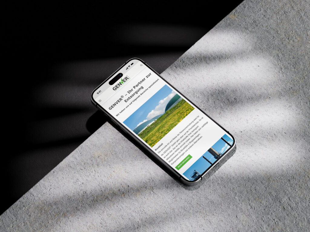 Ein Smartphone, das auf einer strukturierten Oberfläche liegt, zeigt die GENVER-Website mit einem Feld aus grünem Gras, blauem Himmel und weißen Wolken an, wobei der Text und die grüne Schaltfläche von Sonnenlicht und Schatten beleuchtet werden.
