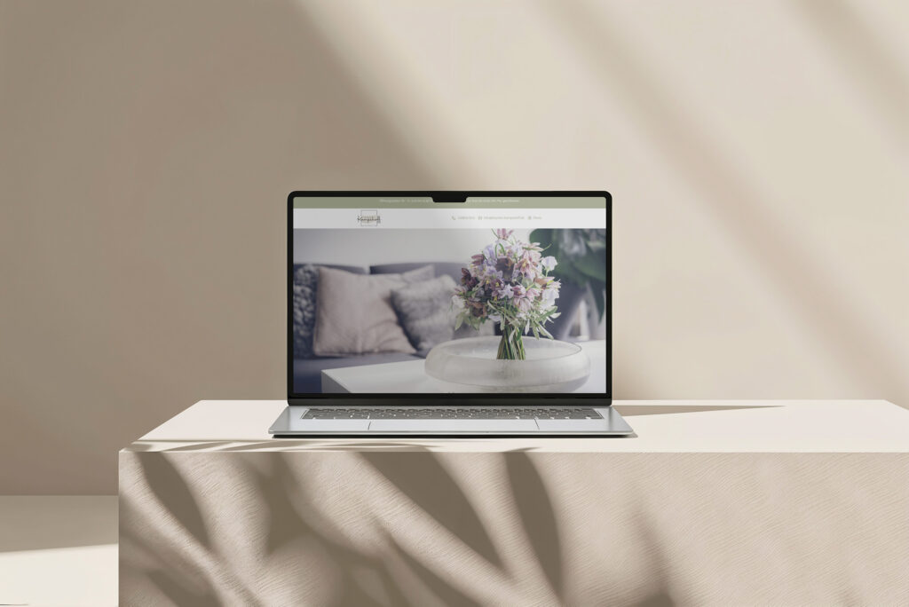 Ein Laptop steht auf einem beigefarbenen Tisch und zeigt die Website des Blumenhauses Kampshoff-van de Sand mit dem Bild einer Blumenvase auf einem Couchtisch in einem modernen, sanft beleuchteten Wohnzimmer.