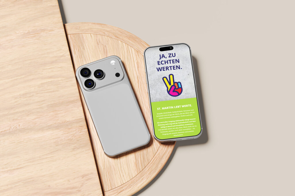 Zwei Smartphones liegen auf einer hellen Holzoberfläche; eines zeigt seine Rückseite mit zwei Kameras, während das andere einen Für Echte Werte One-Pager mit deutschem Text und einem Hand-Friedenszeichen auf dem Bildschirm anzeigt.
