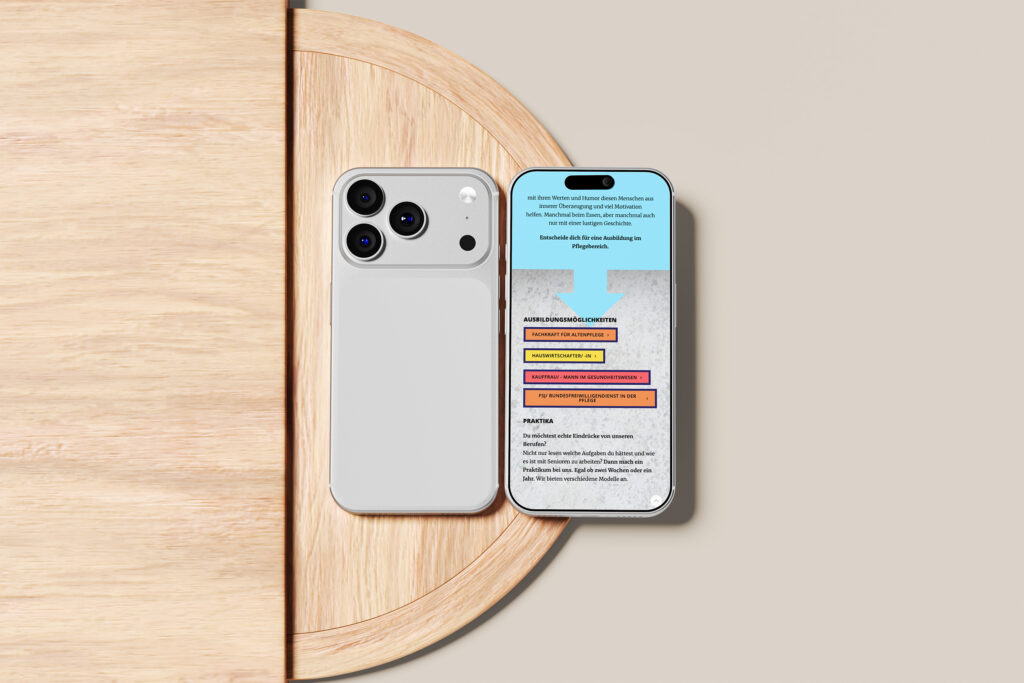 Zwei Smartphones liegen auf einem runden Holztisch; ein Telefon zeigt die Rückseite mit drei Kameras, während das andere die bunte Für Echte Werte One-Pager-Website mit Schaltflächen und deutschem Text auf beigem Hintergrund zeigt.