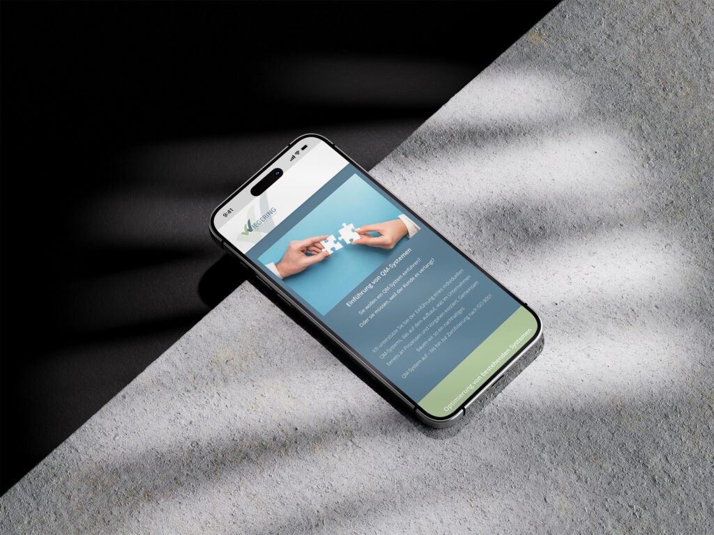 Ein Smartphone liegt auf einer strukturierten Oberfläche im Sonnenlicht und zeigt die Website von Wiegering Beratung & Training mit einem Bild von zwei Händen, die Puzzleteile verbinden, und einem Informationstext darunter an.