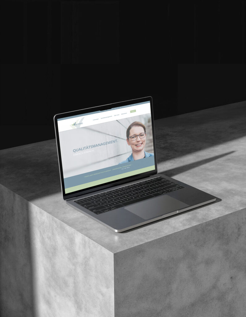 Ein Laptop auf einer Betonfläche zeigt die Website von Wiegering Beratung & Training mit der Überschrift Qualitätsmanagement und dem Foto einer lächelnden Person mit Brille. Die Szene wird von Sonnenlicht mit starken Schatten beleuchtet.