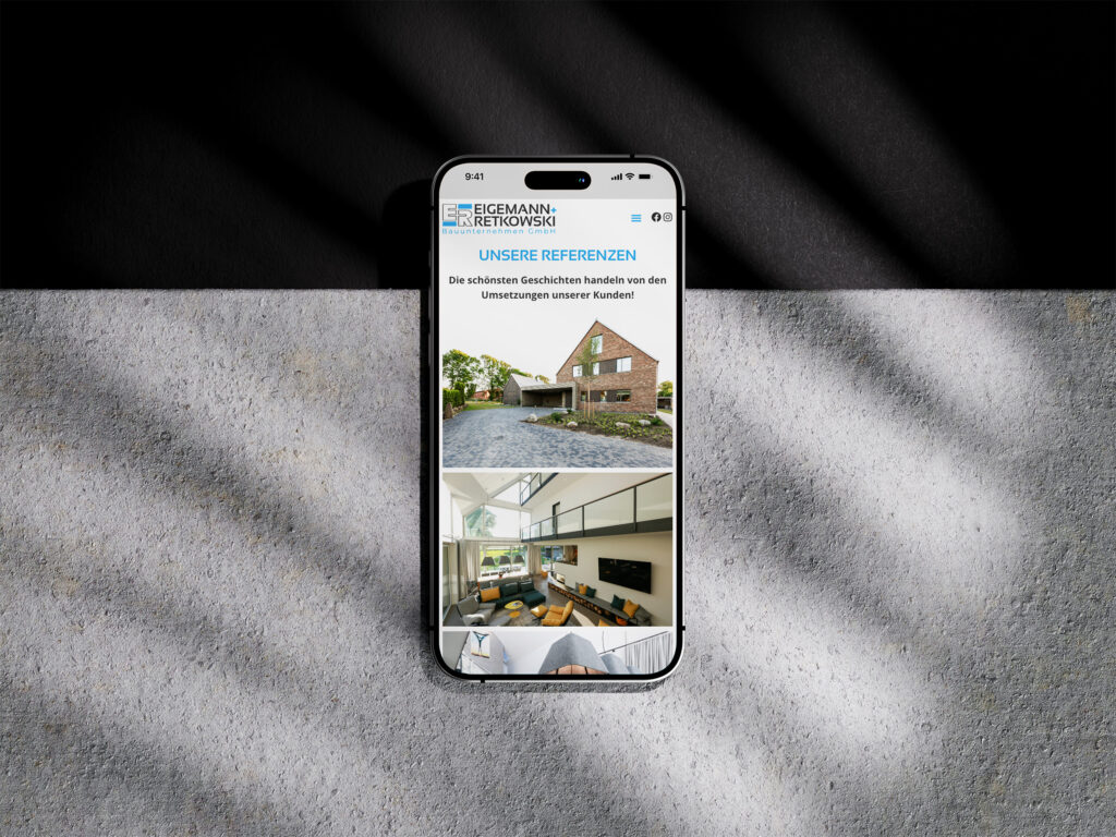 Ein Smartphone zeigt die Eigemann + Retkowski Website Referenz, die ein modernes Haus von außen und eine stilvolle Inneneinrichtung mit deutschem Text unter UNSERE REFERENZEN zeigt. Das Telefon ruht auf einer strukturierten grauen Oberfläche in dramatischem Sonnenlicht und Schatten.