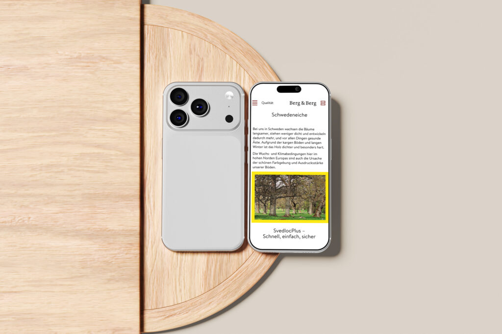 Ein silbernes Smartphone mit einer Dreifachkamera liegt neben einem anderen Telefon, das die Berg & Berg Website Referenz mit Text und einem Foto von Bäumen anzeigt. Beide Telefone liegen auf einem runden Holztisch vor einem beigen Hintergrund.