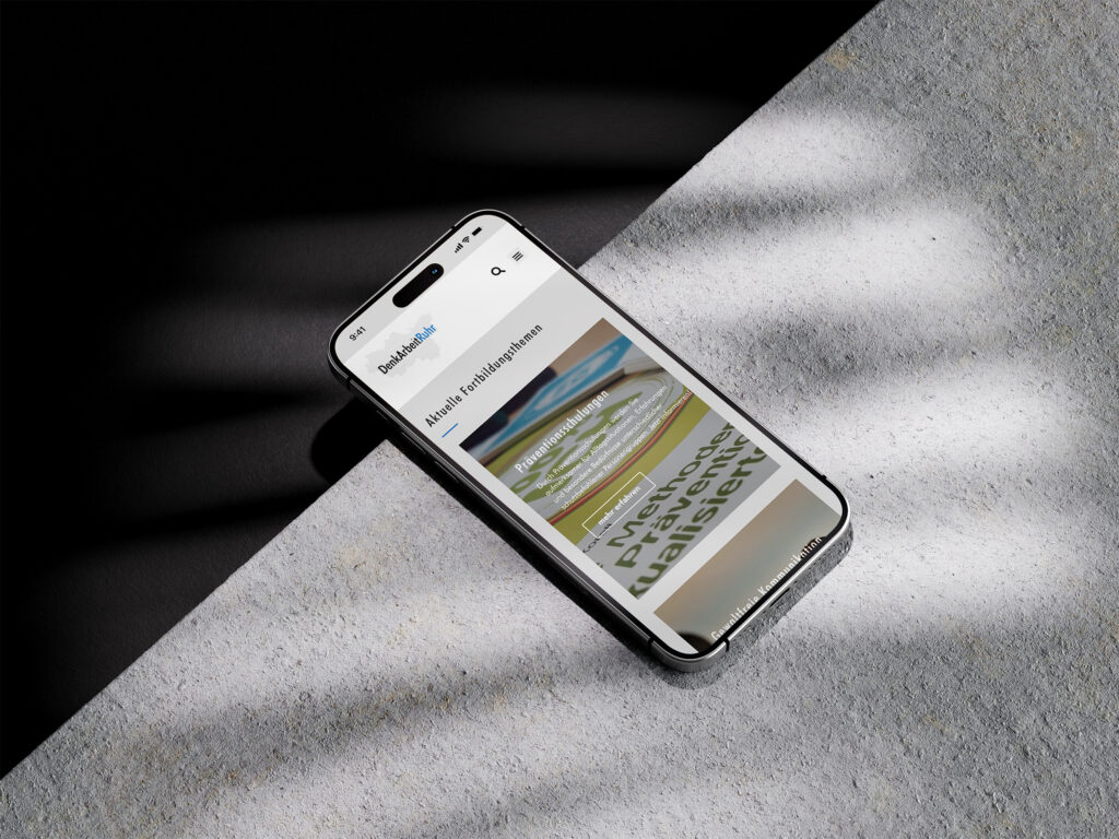 Ein Smartphone liegt auf einer strukturierten grauen Oberfläche im Sonnenlicht und zeigt die DenkArbeit.Ruhr Website mit deutschen Texten und Bildern an. Licht und Schatten erzeugen diagonale Muster in der Szene.