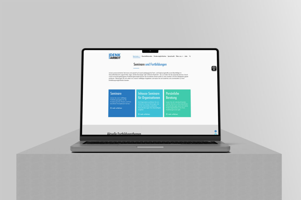 Ein Laptop auf einer grauen Plattform zeigt die DenkArbeit.Ruhr-Website mit weißem Hintergrund, blauen Akzenten und drei farbigen Kästen mit den Bezeichnungen "Seminare", "Inhouse-Seminare für Organisationen" und "Persönliche Beratung".