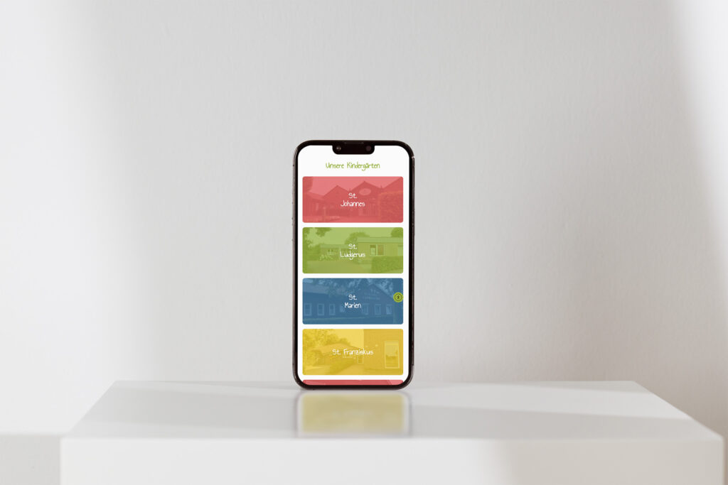 Ein Smartphone steht aufrecht auf einer weißen Fläche und zeigt die App Familienzentrum Heek Website Referenz mit drei bunten Abschnitten, die mit "St. Johannes", "St. Marien" und "St. Franziskus" beschriftet sind, vor einem schlichten hellen Hintergrund.
