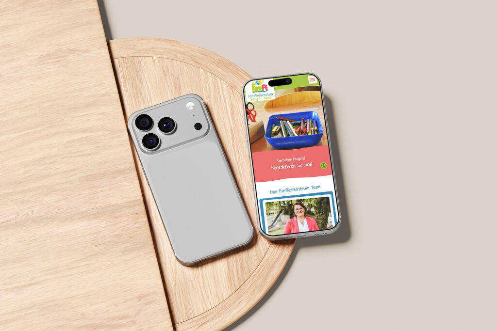 Ein silbernes Smartphone liegt mit der Vorderseite nach unten neben einem anderen Smartphone, das die bunte Familienzentrum Heek Website Referenz anzeigt, auf einem runden Holztisch vor einem beigen Hintergrund.
