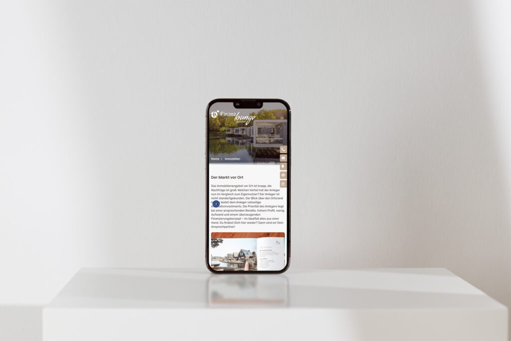 Ein Smartphone steht aufrecht auf einer weißen Fläche vor einer schlichten, hellen Wand und zeigt die Website der Finanzlounge Gescher mit einem Foto eines Hauses am Wasser und einem deutschen Text an.