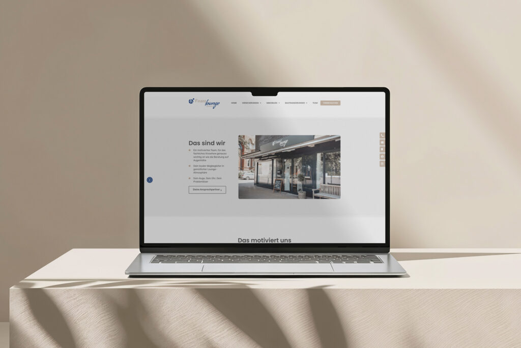 Ein Laptop steht auf einer beigen Fläche und zeigt die Website der Finanzlounge Gescher auf Deutsch mit einem Foto eines Schaufensters. Weiches natürliches Licht wirft Schatten auf die Wand im Hintergrund.