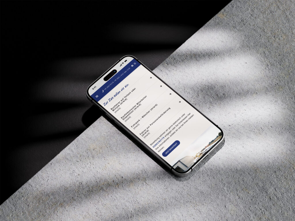 Ein Smartphone liegt auf einer strukturierten Oberfläche und zeigt die Website von Fragemanns Reisekutsche auf Deutsch mit einer weißen und blauen Oberfläche an. Sonnenlicht und Schatten verdecken teilweise sowohl das Telefon als auch die Oberfläche.