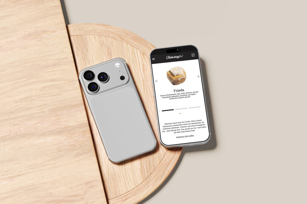 Ein silbernes Smartphone und ein weiteres Telefon mit einer App zur Identifizierung von Kristallen, die beide als Fricke bringt's Website Referenz vorgestellt werden, liegen auf einem hellen runden Holztisch vor einem beigen Hintergrund.