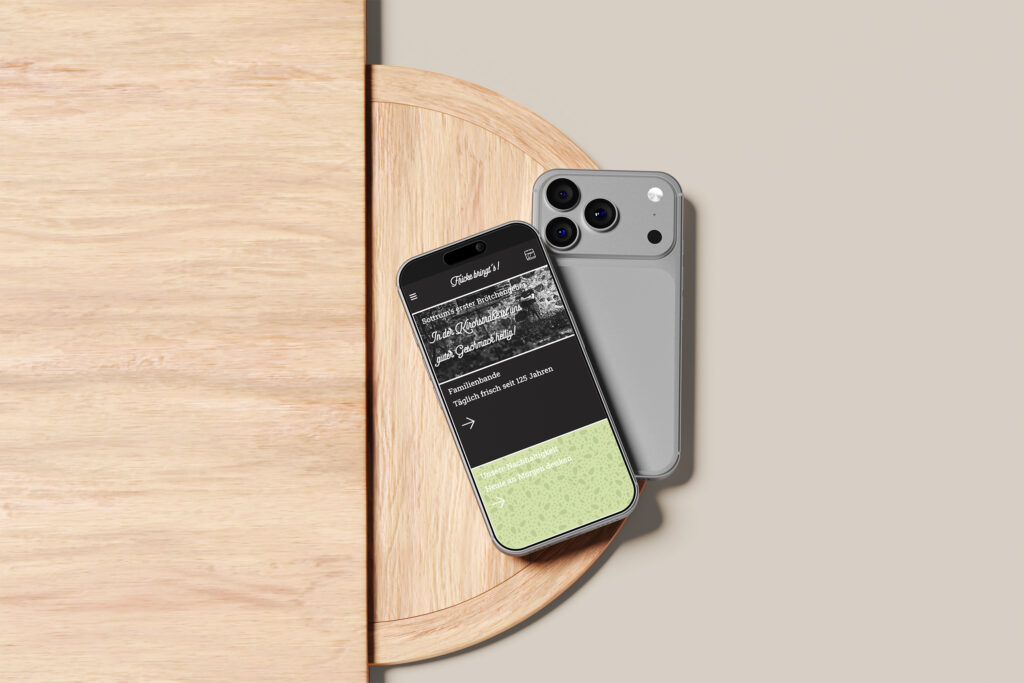 Zwei Smartphones mit silbernen Rückseiten liegen auf einer runden Holzfläche, eines mit der Vorderseite nach oben, auf dem die Fricke bringt's Website Referenz mit schwarzem und grünem Text steht, das andere mit der Rückseite nach unten, alles vor einem hellbeigen Hintergrund.