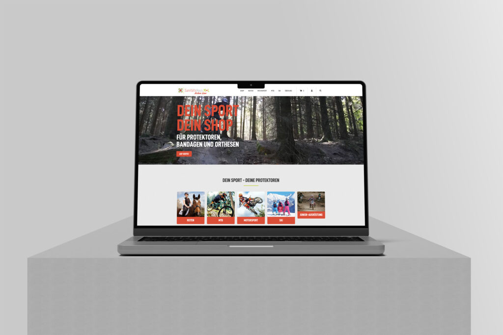 Ein Laptop auf einer grauen Oberfläche zeigt den Sanitätshaus M+L Online-Shop mit einer sportlichen Website mit einem Waldhintergrund, fettgedruckter roter Schrift und vier Kategoriebildern mit den Bezeichnungen Kite, Motorsport, Bike und Dein Geschenk.