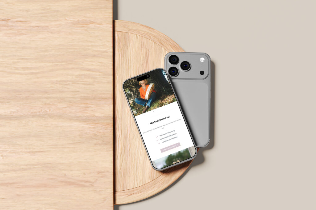 Zwei Smartphones liegen auf einer runden Holzfläche. Ein Telefon zeigt einen Anmeldebildschirm für soziale Medien mit einem Foto einer im Freien sitzenden Frau, das möglicherweise mit der Website von Heike Holdschlag in Verbindung steht, während das andere Telefon nach unten zeigt. Der Hintergrund ist beige.