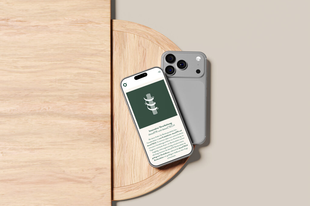 Auf einem runden Holztisch der Dasso Bambus Website liegen zwei Smartphones, eines mit der Vorderseite nach oben, auf dem ein grüner Bildschirm mit Text und einem 3D-Spiralsymbol zu sehen ist, das andere mit der Vorderseite nach unten, auf der die graue Rückseite und die beiden Kameras zu sehen sind.