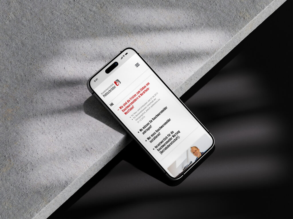 Ein Smartphone, auf dem die Website von Brandschutztechnik Pohlschröder mit deutschem Text angezeigt wird, liegt auf einer hellgrauen Steinoberfläche, die von weichen, schrägen Schatten beleuchtet wird. Der obere Teil des Kopfes einer Person erscheint auf dem Bildschirm des Telefons.