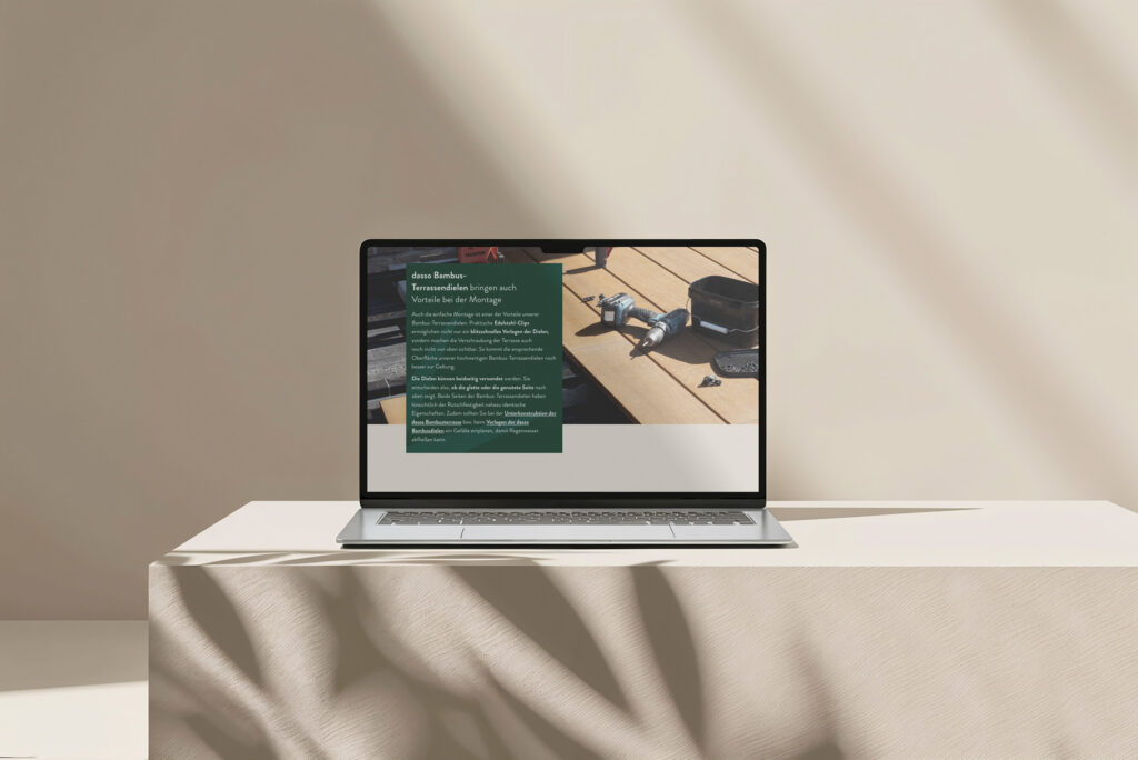 Ein Laptop steht auf einem beigen, stoffbezogenen Tisch im Sonnenlicht. Auf dem Bildschirm erscheint die Website von Dasso Bambus mit deutschem Text und dem Foto einer hölzernen Arbeitsfläche mit Werkzeugen, darunter eine Zwinge und ein Maßband. Der Hintergrund ist schlicht und minimalistisch.