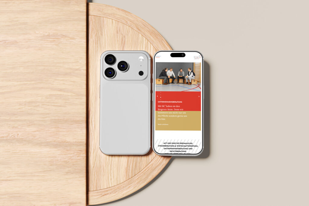 Zwei silberne Smartphones liegen auf einer hellen Holzfläche, eines zeigt seine Rückseite, das andere die SKhoch3-Website mit drei auf einer Bank sitzenden Personen und Text in roten und goldenen Blöcken.