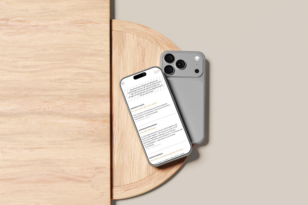 Ein silbernes Smartphone liegt mit der Vorderseite nach unten auf einem runden Holztisch neben einem anderen Telefon, das mit der Vorderseite nach oben liegt und die SKhoch3-Website mit schwarzem und orangefarbenem Text anzeigt. Der Tisch ist teilweise vor einem beigen Hintergrund zu sehen.