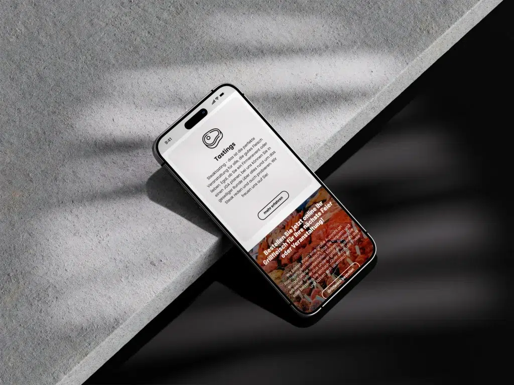 Ein Smartphone liegt auf einer grauen Fläche und zeigt eine App zur Lebensmittelverkostung an, die von der Metzgerei Höing Website Referenz inspiriert wurde. Auf dem Bildschirm werden Informationen zur Verkostung, ein Bild des Lebensmittels und die Schaltfläche "Verkostung buchen" angezeigt, wobei das Sonnenlicht Schatten auf die Szene wirft.