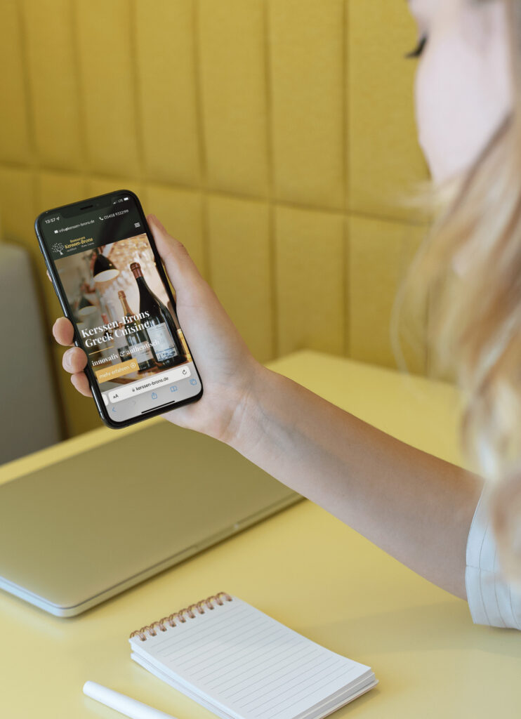 Eine Person hält ein Smartphone in der Hand, auf dem eine Hotelbuchungs-Website angezeigt wird, und sitzt an einem gelben Schreibtisch mit einem geschlossenen Laptop, einem Spiralnotizblock und einem Stift in der Nähe.