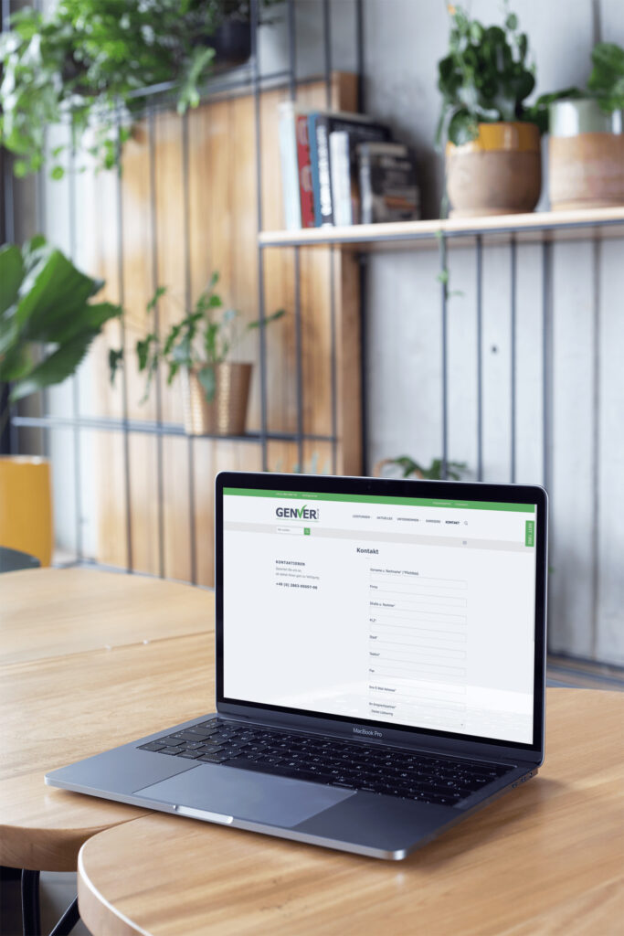 Ein Laptop, auf dem eine Website mit einem Kontaktformular angezeigt wird, steht auf einem runden Holztisch in einem modernen Raum mit Grünpflanzen und Regalen im Hintergrund.