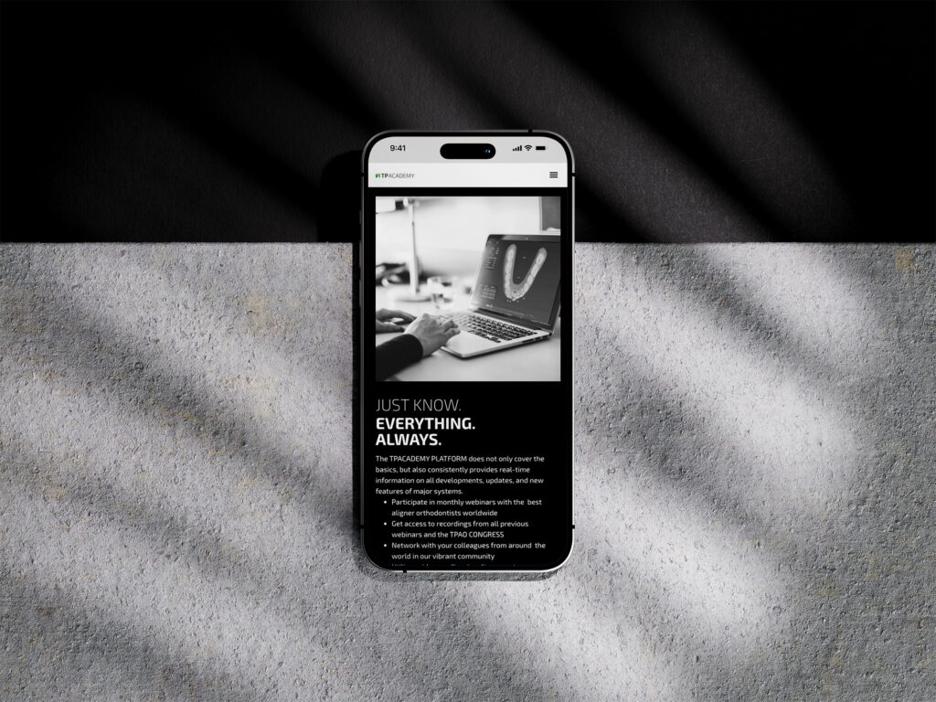 Ein Smartphone zeigt eine monochrome Website der TP Academy mit der Überschrift Just Know. Everything. Always. liegt auf einer strukturierten Oberfläche, über die schräge Schatten geworfen werden. Auf dem Bildschirm sind im Hintergrund eine Tastatur und eine Hand zu sehen.