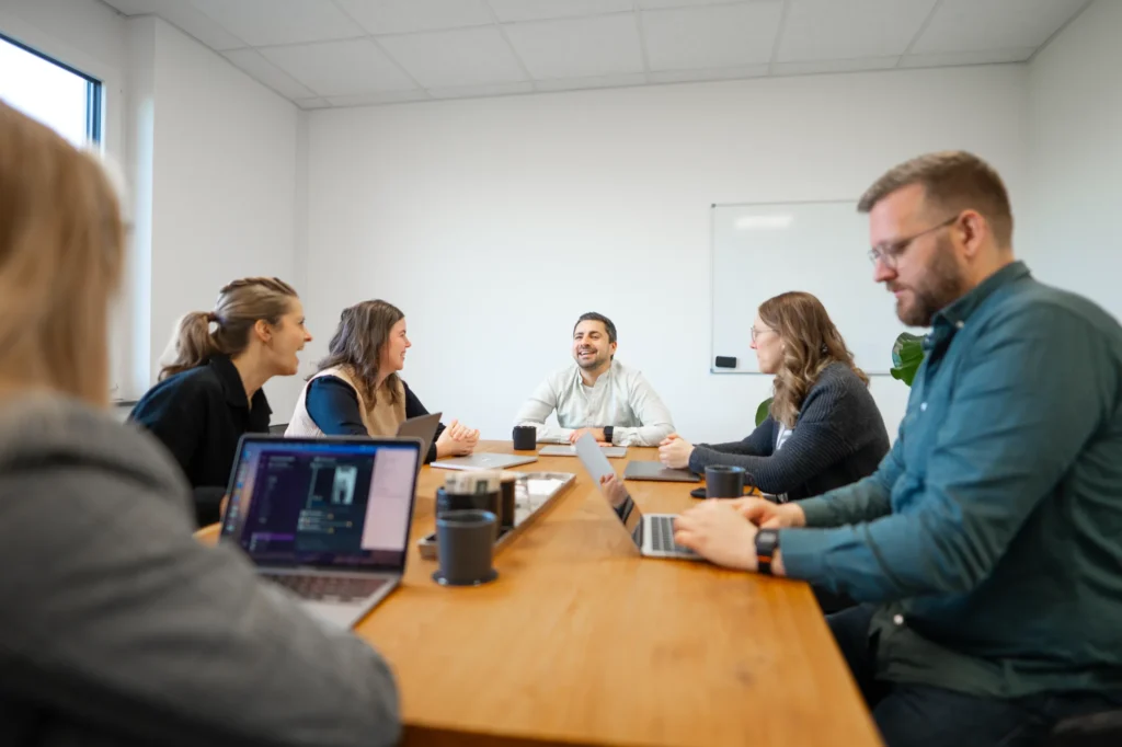 Sechs Personen sitzen um einen hölzernen Konferenztisch in einem hellen Büro und diskutieren. Einige verwenden Laptops, während eine Person das Gespräch leitet. Im Hintergrund sind ein Whiteboard und Fenster zu sehen.