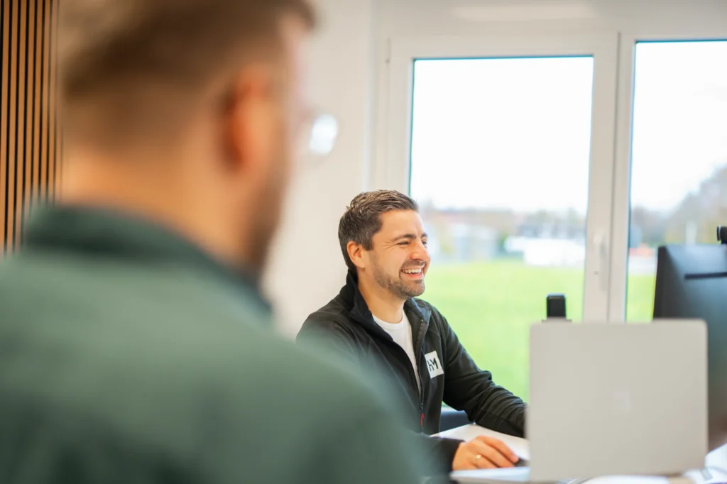 Ein Mann sitzt an einem Schreibtisch und lächelt auf einen Computerbildschirm in einem hellen Büro, vielleicht beschäftigt mit einem Strategiegespräch Online-Marketing, mit einer anderen Person im Vordergrund verschwommen. Große Fenster zeigen ein grünes Feld draußen.