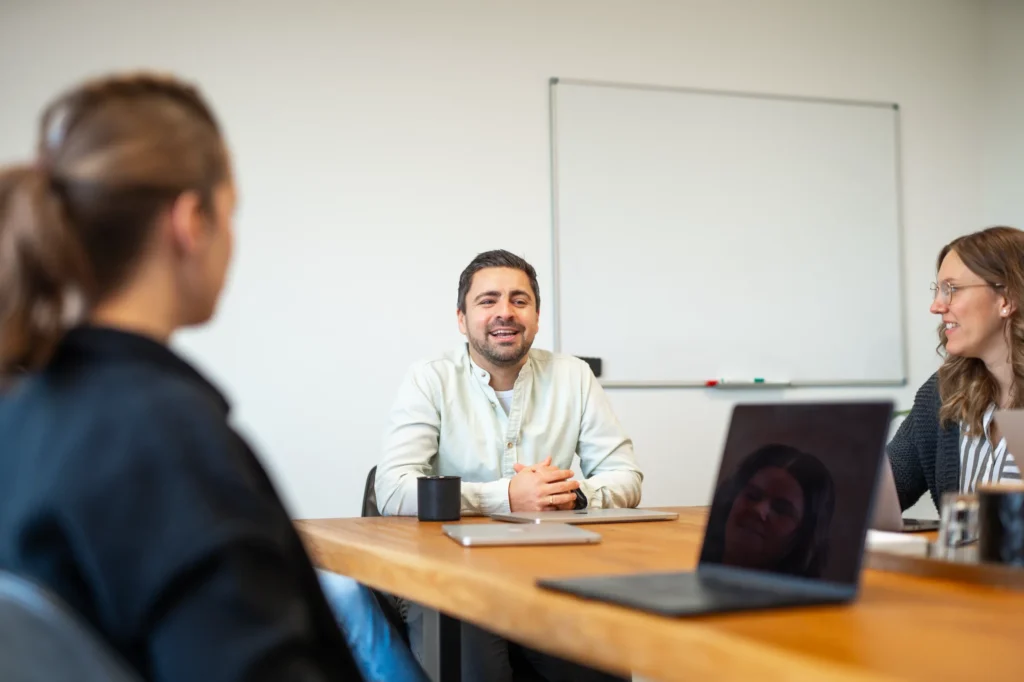 Drei Personen sitzen an einem Konferenztisch und führen eine Diskussion. Eine Person lächelt und spricht, eine andere hört zu, und das Gesicht einer dritten Person spiegelt sich auf einem Laptop-Bildschirm. Im Hintergrund befindet sich ein leeres Whiteboard.