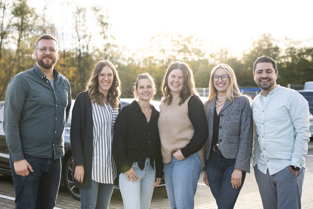 Sechs Erwachsene stehen in legerer Kleidung im Freien und lächeln in die Kamera. Im Hintergrund sind Bäume und geparkte Autos zu sehen. Das Sonnenlicht schafft eine helle, warme Atmosphäre - und fängt den freundlichen Teamgeist der Digitalagentur Münsterland ein.