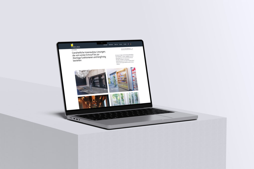 Ein Laptop steht auf einem weißen Sockel und zeigt eine Website mit deutschem Text, einer Kopfzeile und Bildern von verschiedenen Gebäuden und Innenräumen in einem Rasterlayout. Der Bildschirm hat ein klares, modernes Design.