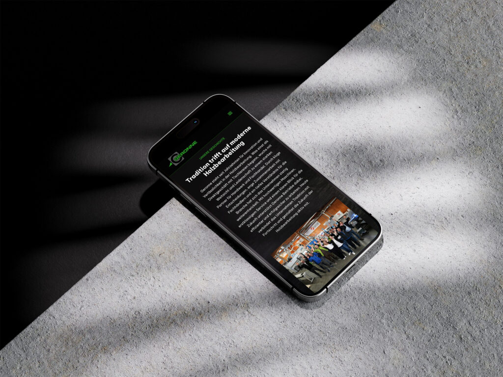Ein Smartphone liegt auf einer strukturierten grauen Oberfläche und zeigt einen Website-Artikel mit Text und einem Gruppenfoto am unteren Rand an. Licht- und Schattenmuster fallen über die Oberfläche und den Bildschirm des Smartphones.