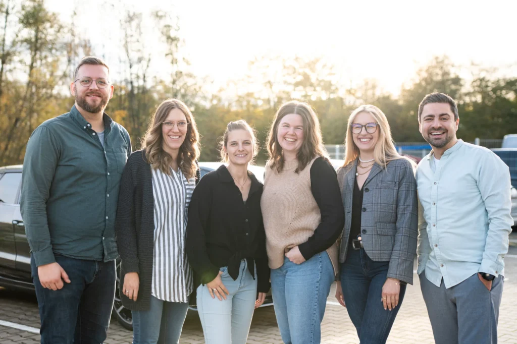 Sechs Erwachsene stehen im Freien auf einem Parkplatz und lächeln in die Kamera. Sie sehen aus wie ein freundliches Team von einer Webagentur für Webseiten & Online-Marketing, das das Sonnenlicht genießt, während im Hintergrund Bäume und Autos zu sehen sind.