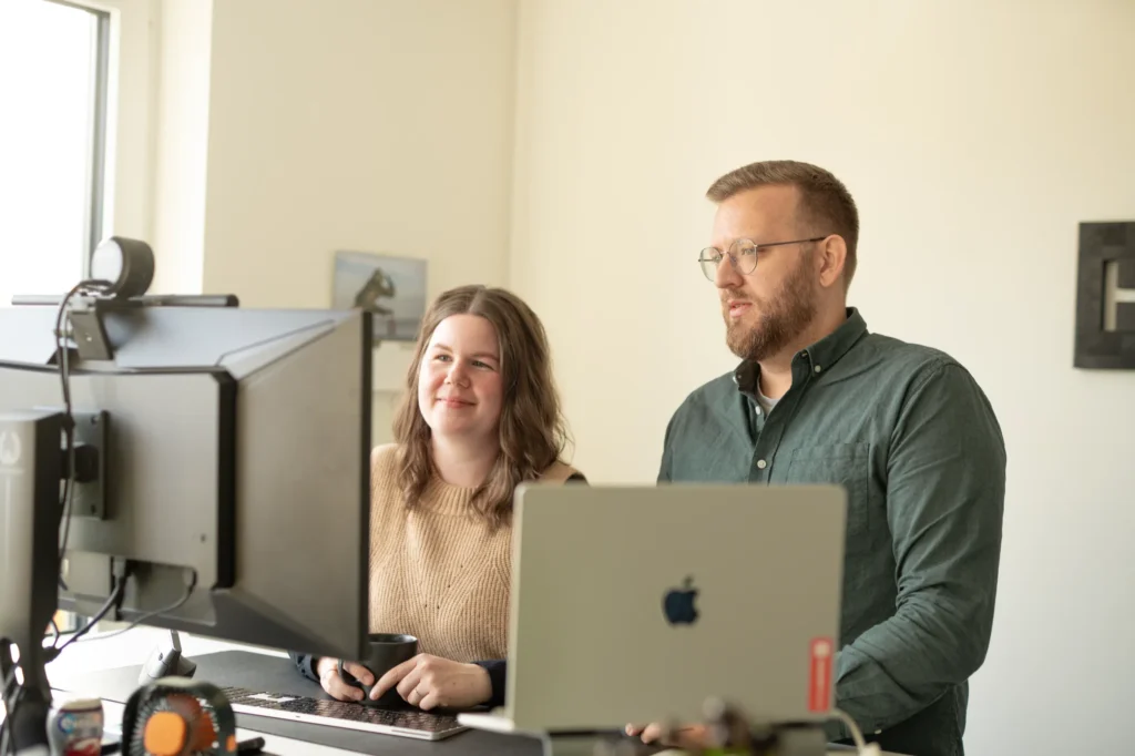 Zwei Personen stehen an einem Schreibtisch und schauen auf Computerbildschirme. Einer hält eine Tasse in der Hand und lächelt, während der andere konzentriert an der Mitarbeitergewinnung zu arbeiten scheint. Ein Laptop und Büroutensilien stehen in der Nähe, Sonnenlicht fällt durch das Fenster.