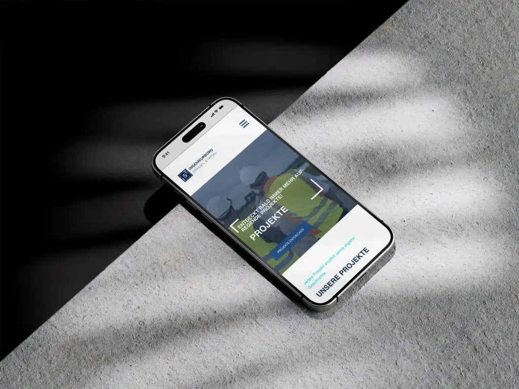 Ein Smartphone, das eine Website mit deutschem Text anzeigt, liegt auf einer strukturierten Oberfläche, die teilweise von Sonnenlicht und Schatten beleuchtet wird. Auf dem Bildschirm sind Bauarbeiter und das Wort PROJEKTE zu sehen.