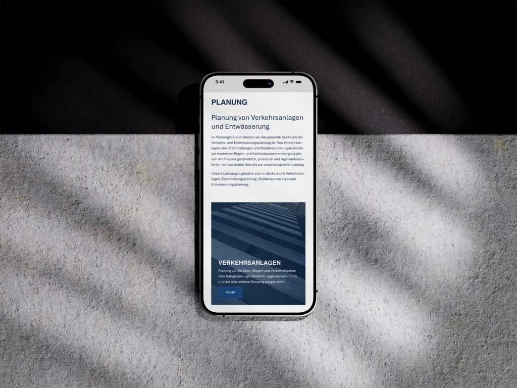 Ein Smartphone, das eine deutschsprachige Website über die Planung von Verkehrssystemen anzeigt, liegt auf einer strukturierten Oberfläche mit dramatischen Licht- und Schattenmustern. Der Bildschirm zeigt Text und das Bild einer Straße mit Zebrastreifen.
