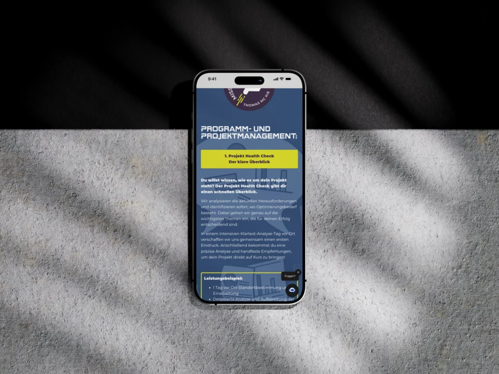 Ein Smartphone liegt auf einer grauen Fläche, auf die Schatten geworfen werden. Auf dem Bildschirm erscheint eine Website über Programm- und Projektmanagement in deutscher Sprache mit blauem Hintergrund und weißem und gelbem Text.