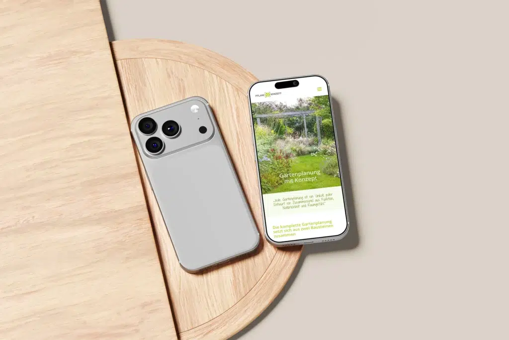 Zwei moderne Smartphones sind auf einem hellen runden Holztisch platziert. Ein Telefon zeigt eine Website zur Gartenplanung an, während das andere Telefon mit der Vorderseite nach unten liegt und seine Kameralinsen zeigt.