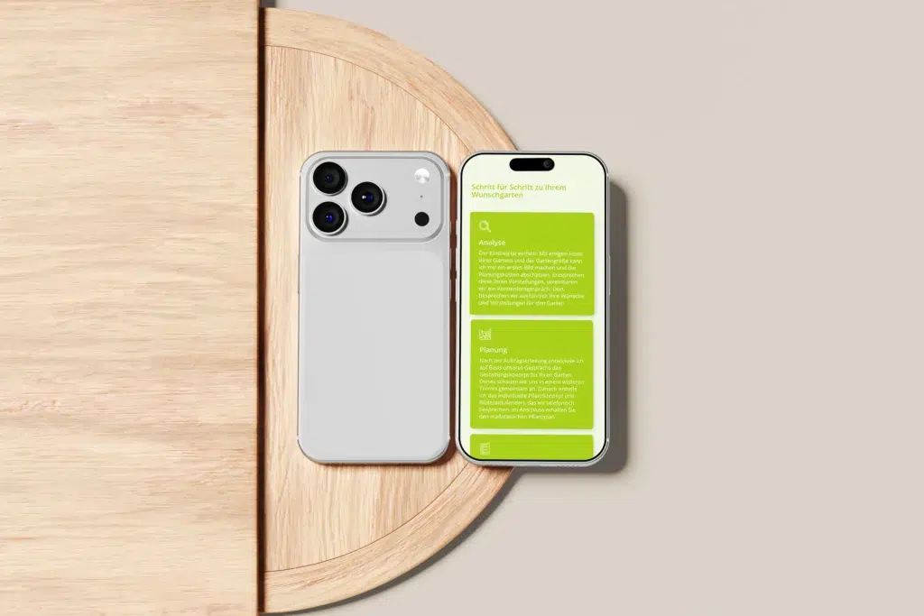 Ein weißes Smartphone mit drei rückwärtigen Kameras liegt auf einem runden Holztisch neben einem anderen Telefon, auf dem ein grüner App-Bildschirm mit deutschen Texten und Symbolen angezeigt wird, alles vor einem hellbeigen Hintergrund.