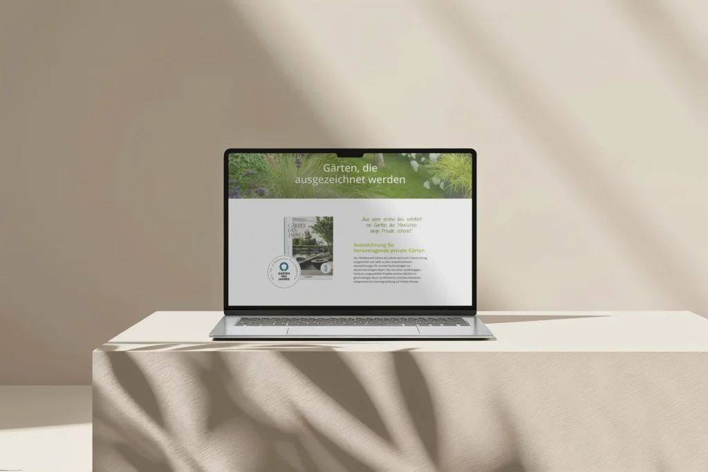 Ein Laptop auf einem beigen Tisch zeigt eine deutschsprachige Webseite über preisgekrönte Gärten mit Text, einem Foto eines Gartenbuchs und einem Pflanzenhintergrund. Das Sonnenlicht wirft Schatten auf die Szene.