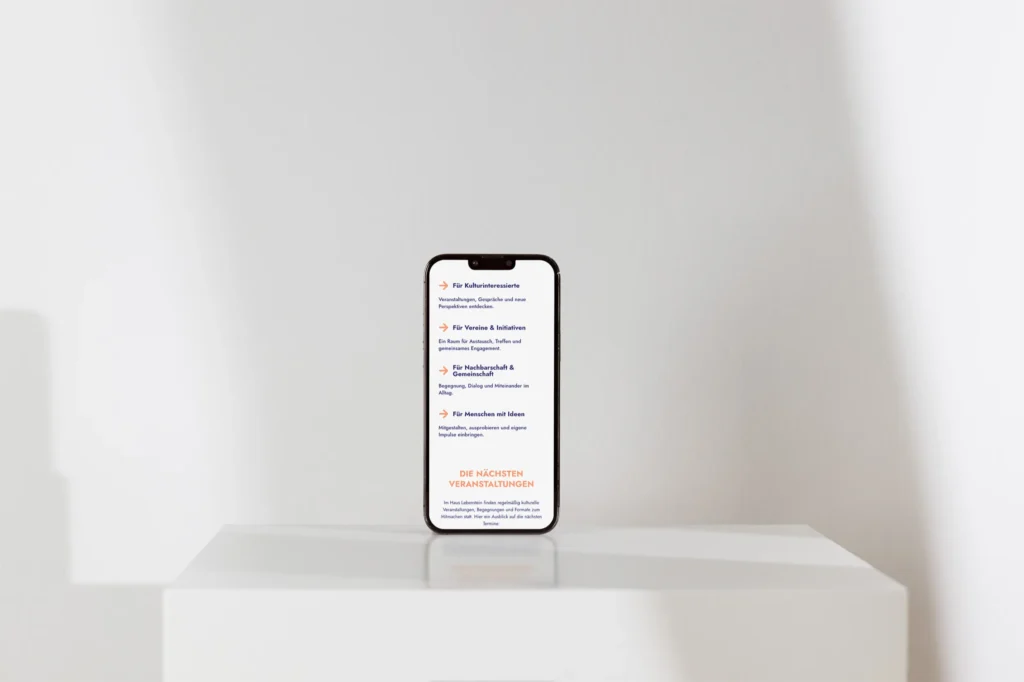 Ein Smartphone steht aufrecht auf einer weißen Fläche und zeigt eine Liste mit Text in deutscher Sprache an. Der Hintergrund ist schlicht weiß und weist sanfte Schatten auf.