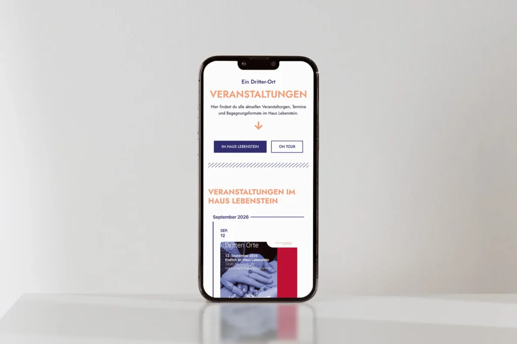 Auf einem Smartphone wird eine Website auf Deutsch über bevorstehende Veranstaltungen im Haus Lebenstein angezeigt, auf der Veranstaltungstermine, Schaltflächen und ein Bild in Blautönen zu sehen sind. Das Smartphone steht aufrecht auf einer weißen Fläche vor einem hellgrauen Hintergrund.