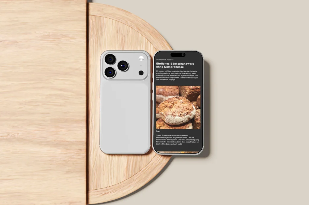 Ein weißes Smartphone liegt mit dem Display nach unten neben einem anderen, auf dem eine deutsche Webseite über frisch gebackenes Brot zu sehen ist, auf einem runden Holztisch vor einem beigen Hintergrund.