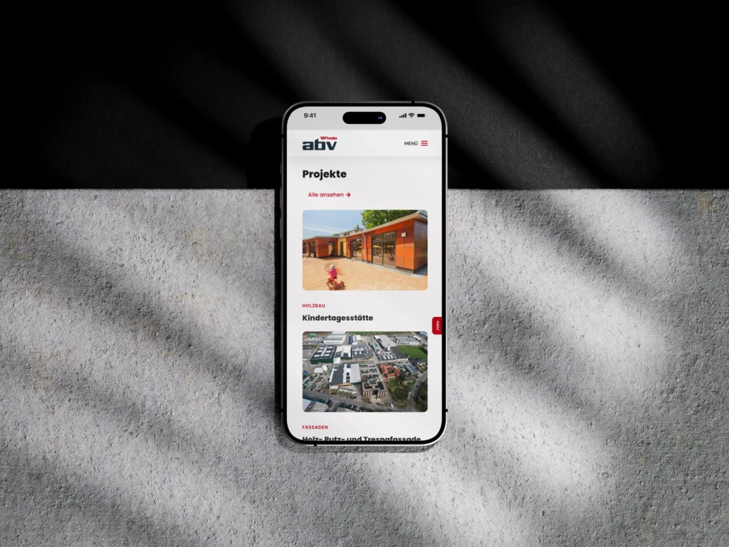 Auf einem Smartphone wird eine Website mit Architekturprojekten angezeigt, darunter auch ein Kindergarten. Das Smartphone liegt auf einer strukturierten Oberfläche, über die diagonale Licht- und Schattenmuster verlaufen.