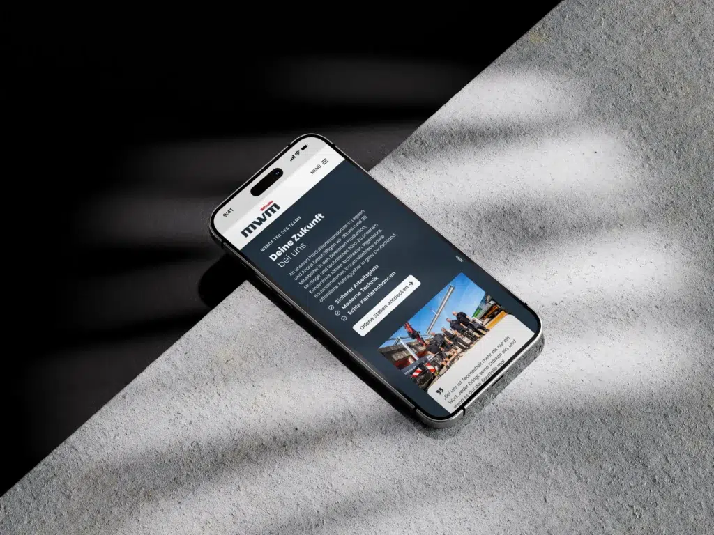 Ein Smartphone mit dunklem Bildschirm, auf dem eine Website mit dem Titel „MVMT“ zu sehen ist, liegt auf einer strukturierten, hellen Oberfläche mit diagonalen Schatten. Die Website zeigt Text und ein Bild von Menschen auf einer Baustelle in der Nähe von Kränen.