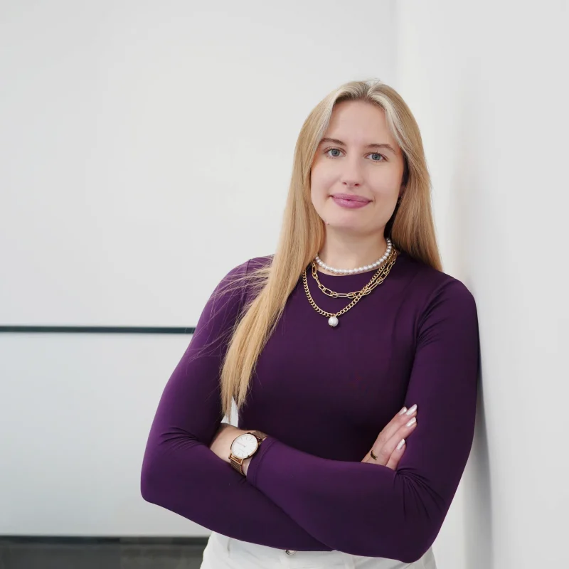 Eine Frau mit langen blonden Haaren, die ein lilafarbenes langärmeliges Oberteil, mehrere Ketten und eine Uhr trägt, steht mit verschränkten Armen und einem leichten Lächeln vor einer weißen Wand in einer modernen, hellen Umgebung.