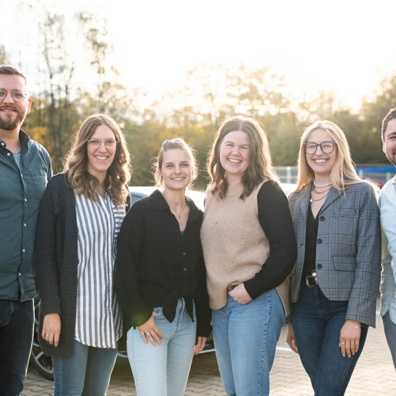 Sechs Erwachsene stehen im Freien auf einem Parkplatz und lächeln in die Kamera. Sie sehen aus wie ein freundliches Team von einer Webagentur für Webseiten & Online-Marketing, das das Sonnenlicht genießt, während im Hintergrund Bäume und Autos zu sehen sind.