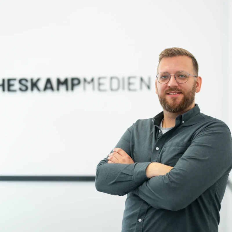 Ein Mann mit Brille und Bart steht lächelnd mit verschränkten Armen vor einem unscharfen Schild mit der Aufschrift Heskamp Medien an einer weißen Wand, das für Jaron Heskamp Digitale Visitenkarte steht.